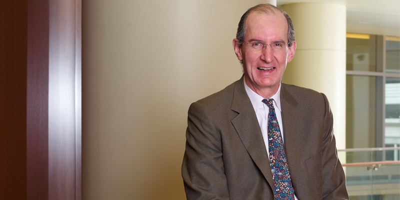 Wearing a brown suit with a dark blue, brown, and burgundy tie, Dr. Thomas Mustoe stands in front of an office wall smiling.