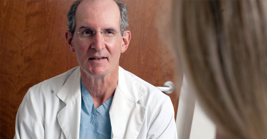 Dr. Thomas Mustoe wears a white lab coat and light blue scrubs while consulting with a blond patient.