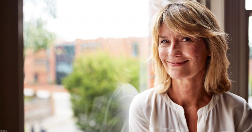 Smiling blond woman in white blouse sits in front of a window overlooking a brick building and trees in a campus-like setting.