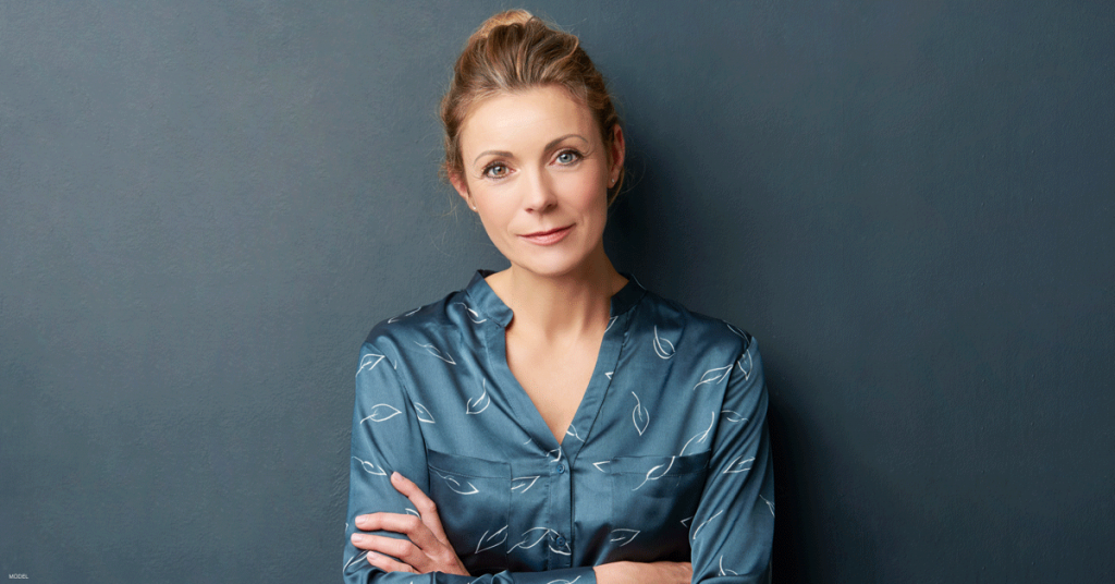 Woman in teal blue blouse crosses her arms and leans her back against a dark blue wall.