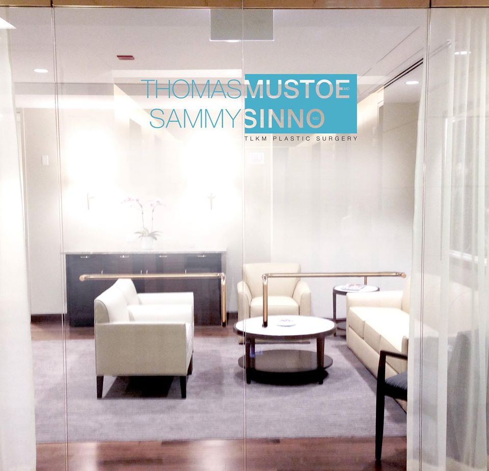 Glass doors open onto TLKM Plastic Surgery's reception area that has modern white sofas and armchairs and dark wood accents. 