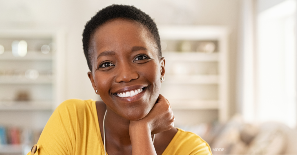 A black women model with short hair smiling with her hand resting under her chin, and she is wearing a yellow shirt.
