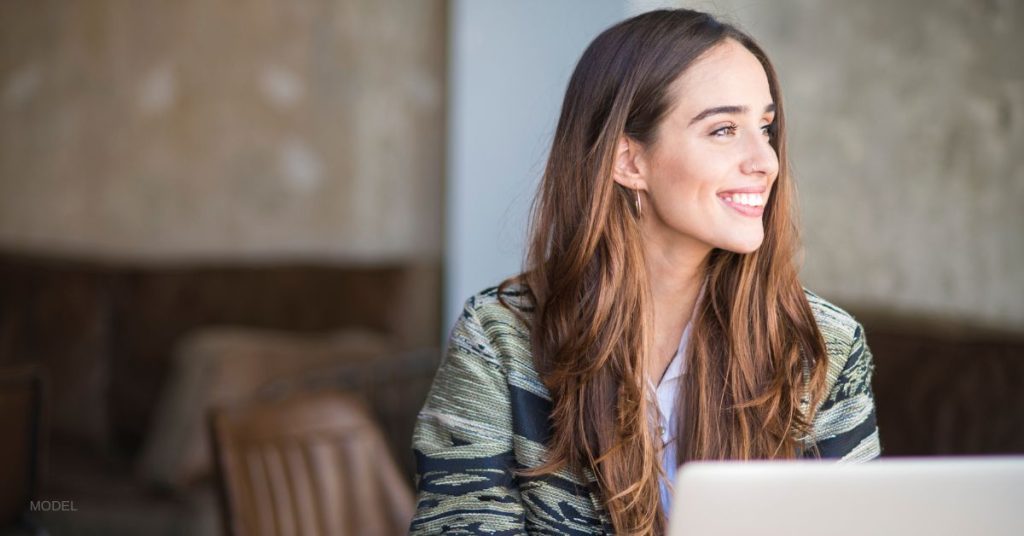 Beautiful woman sits a computer and smiles while looking out the window. (Model)