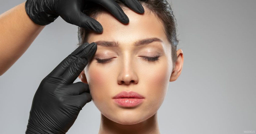 A woman getting their eyebrows examined by someone with gloves. (Models)